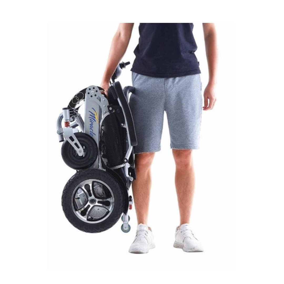 A person wearing a black t-shirt, gray shorts, and white sneakers stands holding a folded Miracle Mobility Platinum 8000 Bariatric Portable Powerchair by its frame. The compact electric wheelchair features large wheels.