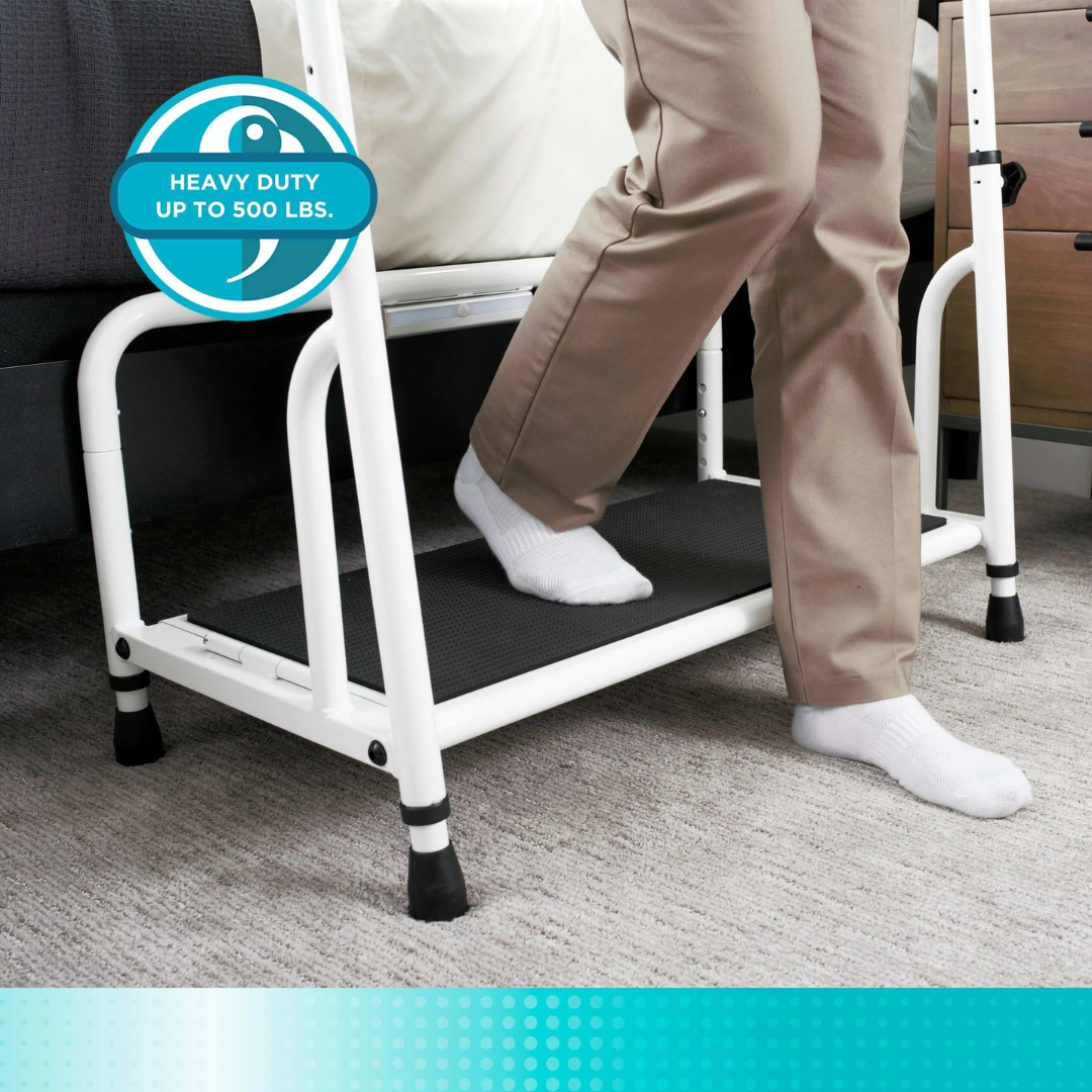 A person in beige pants and white socks steps onto the Stander Step Bedside Platform with LED Lighting, featuring black non-slip surfaces and a white frame for safety. The image notes it is heavy duty, supporting up to 500 lbs.