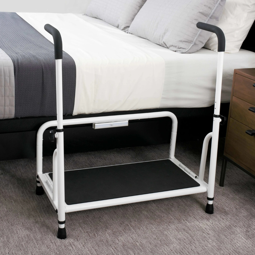 The Stander Step Bedside Platform with LED Lighting, featuring sturdy white metal construction, black handles, a non-slip surface, and motion-activated LED light, is placed beside a bed near a wooden nightstand on grey carpet.