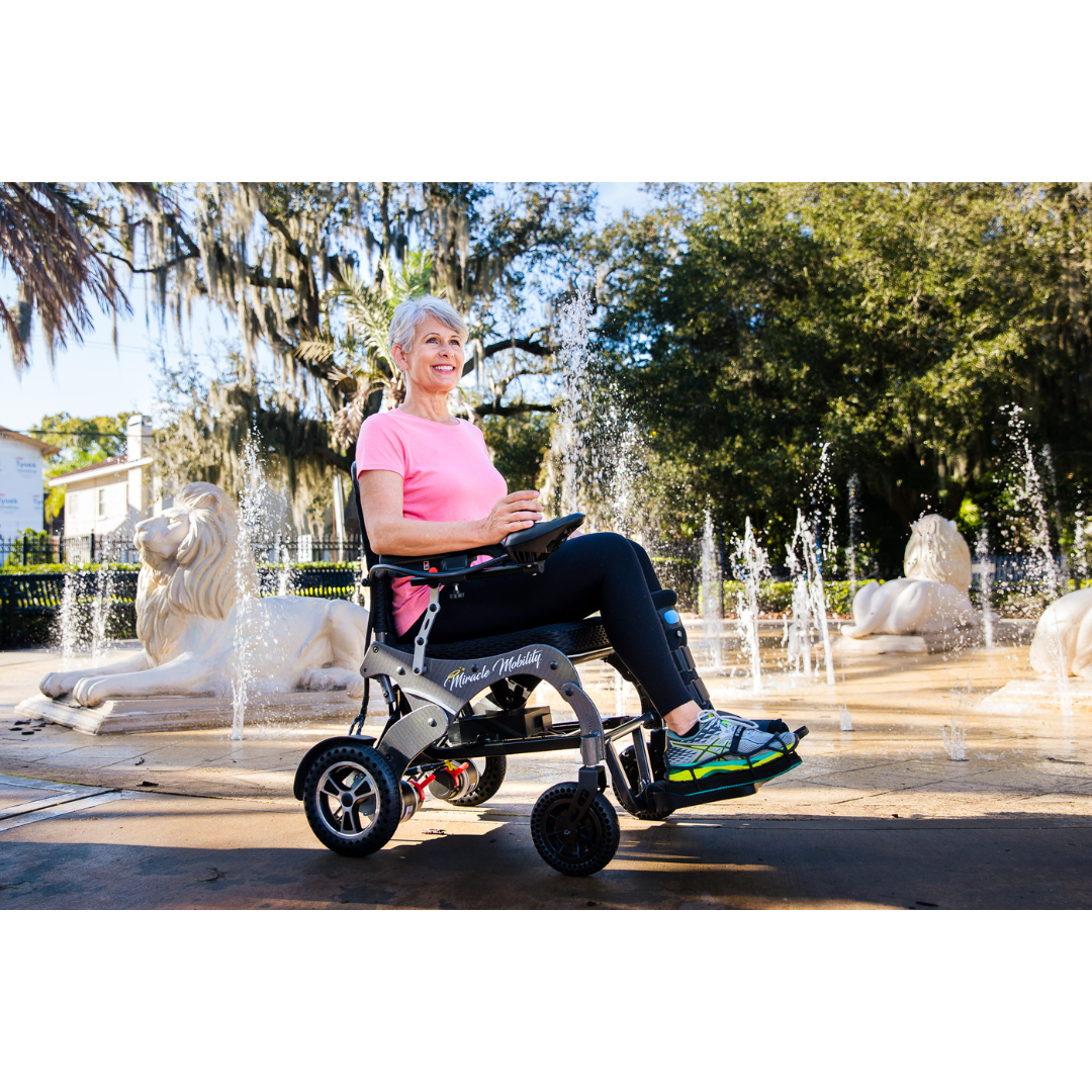A smiling older woman in a pink shirt sits in a Miracle Mobility Palladium 8500 Portable Powerchair near a fountain with water jets and white lion statues, surrounded by greenery and sunlight.