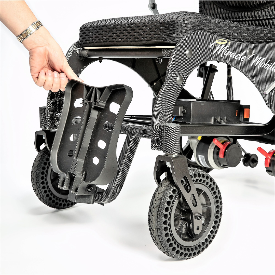 A hand is adjusting or removing the footrest of the Miracle Mobility Palladium 8500 Portable Powerchair, a lightweight, foldable wheelchair with black wheels and a textured frame.
