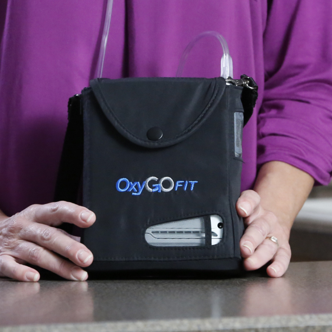 A person in a purple shirt holds a lightweight OxyGo FIT Compact Portable Oxygen Concentrator in a black carrying case on a countertop.