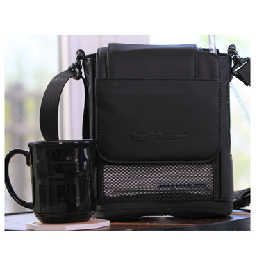 A black mug sits beside an OxyGo NEXT Carry Bag with Shoulder Strap, which features a mesh pocket for heat ventilation and an adjustable storage strap, displayed on a table near a bright window.