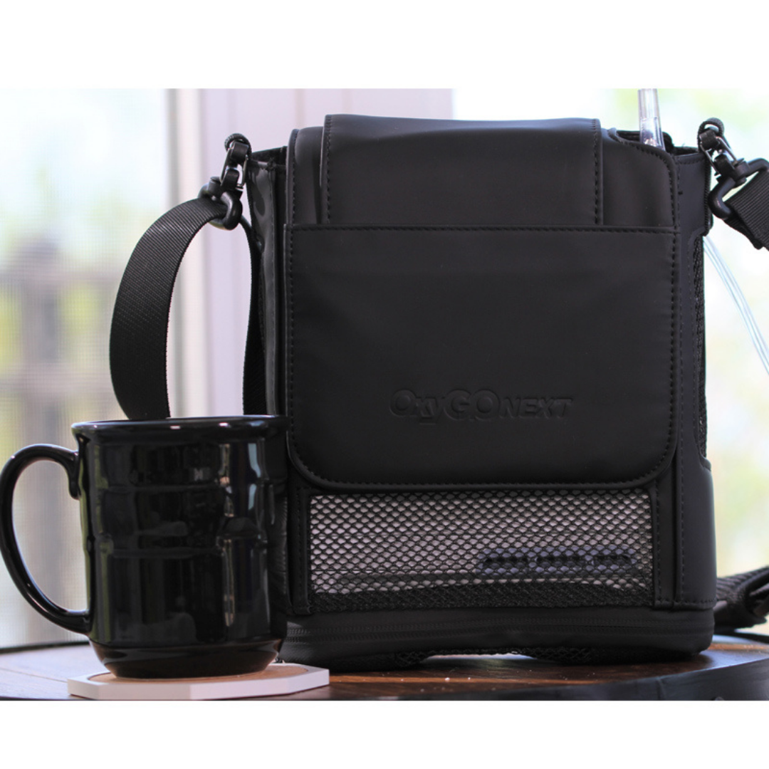 An OxyGo NEXT Ultralite Portable Oxygen Concentrator black shoulder bag rests on a table beside a black ceramic mug, with natural light streaming through a window in the softly blurred indoor background.
