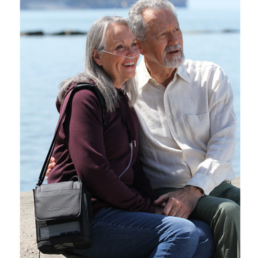 An older woman enjoys a sunny day by the water with an older man, smiling and carrying her OxyGo NEXT Ultralite Portable Oxygen Concentrator over her shoulder.
