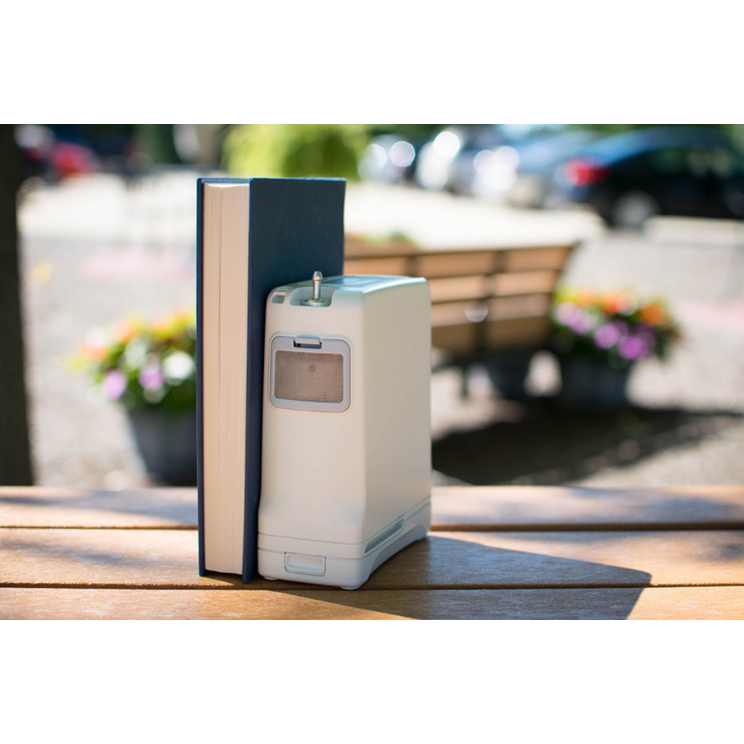 A medical device, like the OxyGo FIT Compact Portable Oxygen Concentrator, and a hardcover book stand upright side by side on a wooden table outdoors, with blurred flowers, benches, and parked cars in the background.