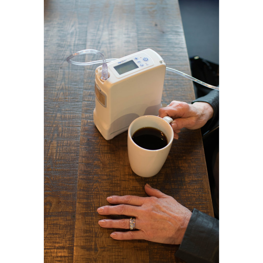 A person with a ring on their finger holds a white coffee mug on a wooden table beside an OxyGo FIT Compact Portable Oxygen Concentrator with a nasal cannula tube.