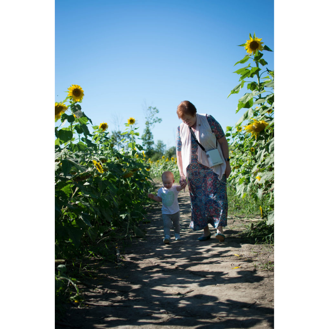 A woman carries her OxyGo FIT Compact Portable Oxygen Concentrator as she and a small child walk hand-in-hand along a dirt path lined with sunflowers under a clear sky. She wears a long dress and scarf; the child, a light outfit.