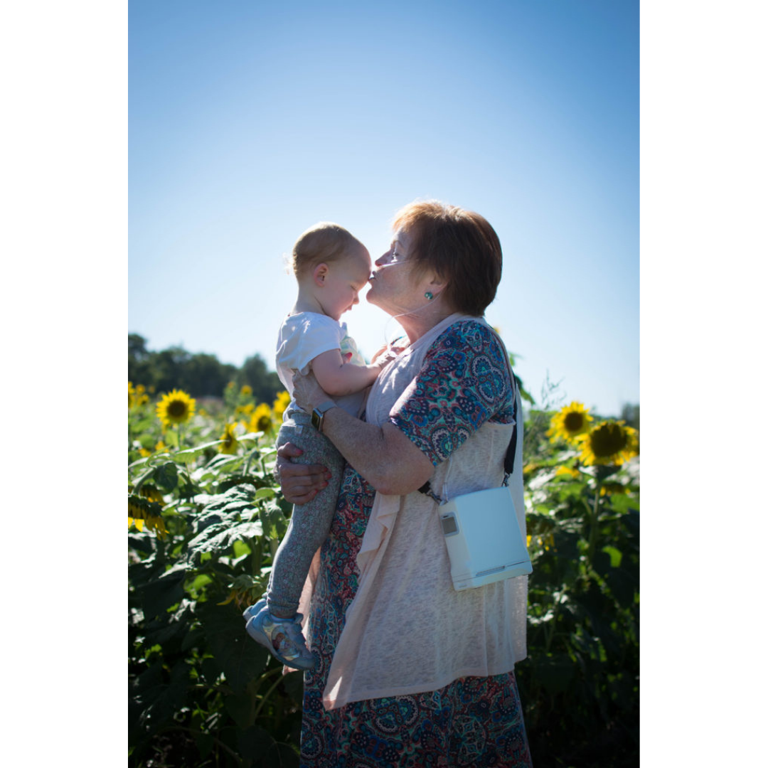 An older woman smiles and holds a baby in a sunny sunflower field, her OxyGo FIT Compact Portable Oxygen Concentrator discreet beside her. They touch foreheads affectionately, happy together with blue sky and sunflowers behind them.