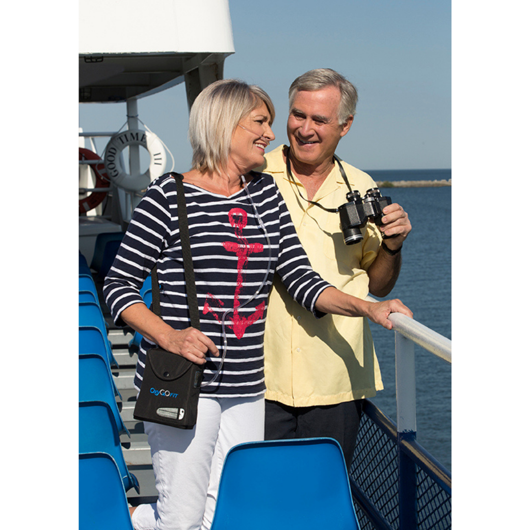 A smiling older couple stands on a boat deck by the water; the woman wears a striped shirt with a red anchor and carries a black bag, while the man holds binoculars and their lightweight OxyGo FIT Compact Portable Oxygen Concentrator.