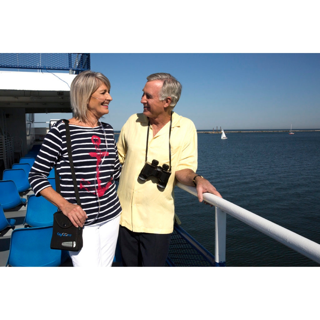 An older couple smiles at each other on a ferry deck, enjoying blue skies and distant sailboats. The man wears binoculars and carries an OxyGo FIT Compact Portable Oxygen Concentrator, making travel easy and relaxing.