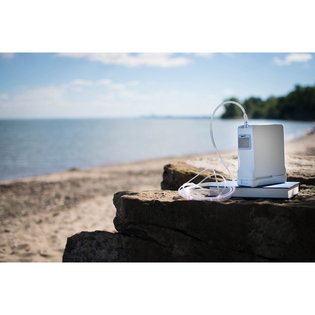The OxyGo FIT Compact Portable Oxygen Concentrator with attached tubing rests on a rock by a sandy beach, providing mobile oxygen therapy near calm water and distant trees under a partly cloudy sky.