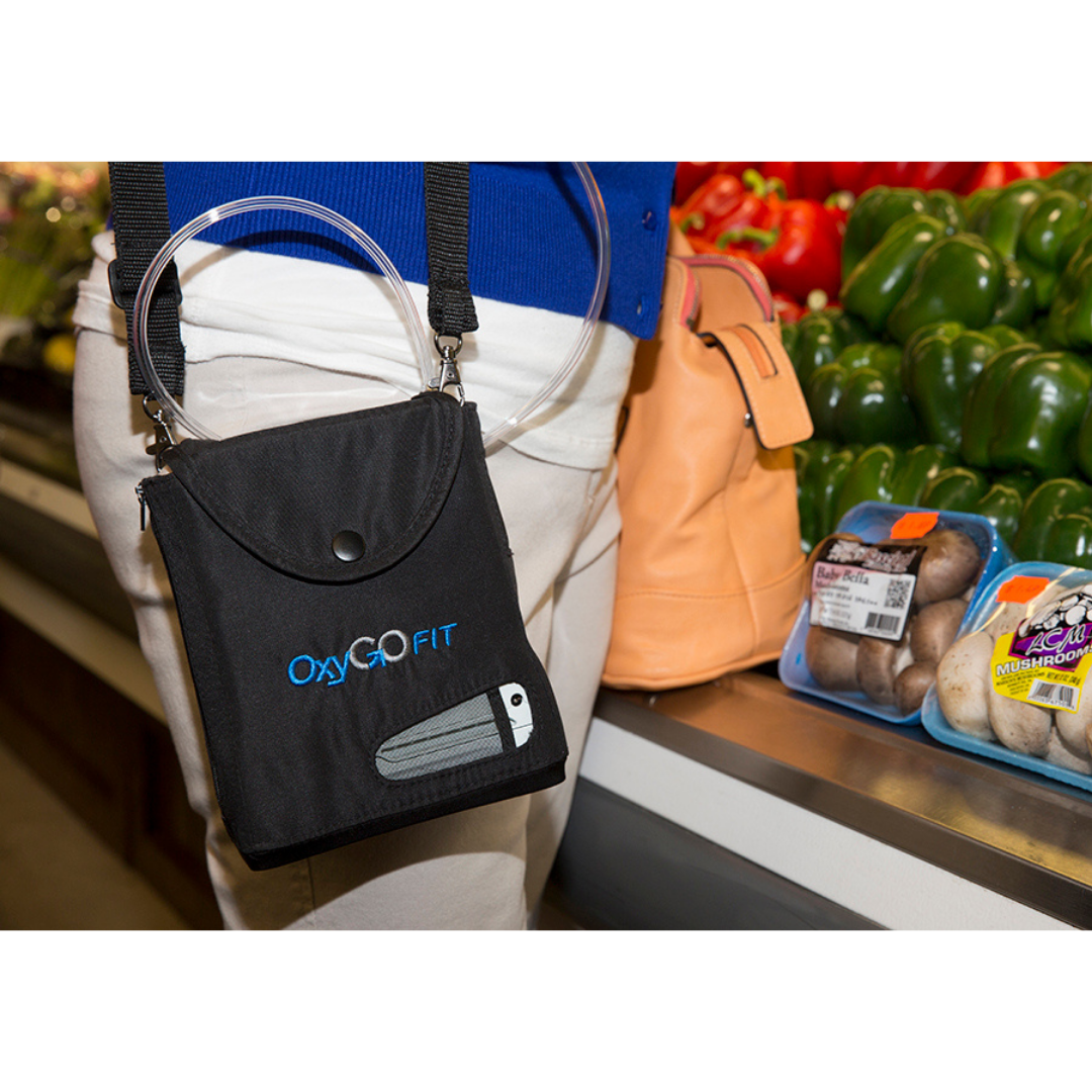 A person stands in a grocery store produce section, carrying an OxyGo FIT Compact Portable Oxygen Concentrator in a shoulder bag. Fresh bell peppers and packaged mushrooms are visible in the background.