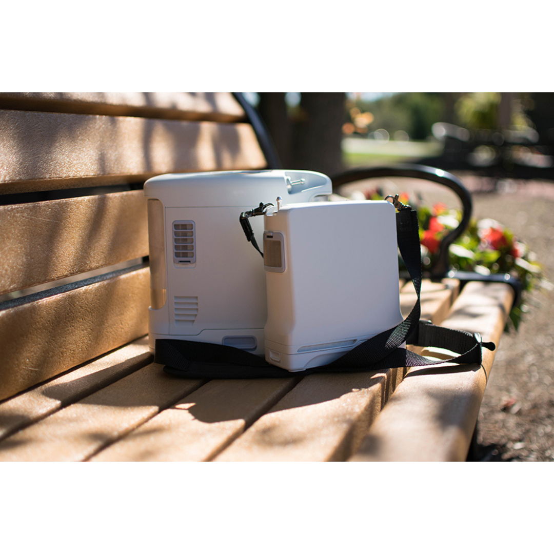 Two OxyGo FIT Compact Portable Oxygen Concentrators with shoulder straps rest on a sunlit wooden park bench, with blurred greenery and flowers in the background, showcasing the convenience of portable oxygen therapy.