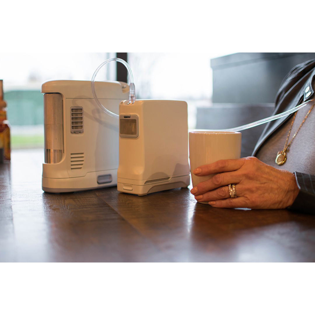 A person holds a white mug with one hand while using the OxyGo FIT Compact Portable Oxygen Concentrator on a wooden table. A clear tube connects the concentrator to the person.