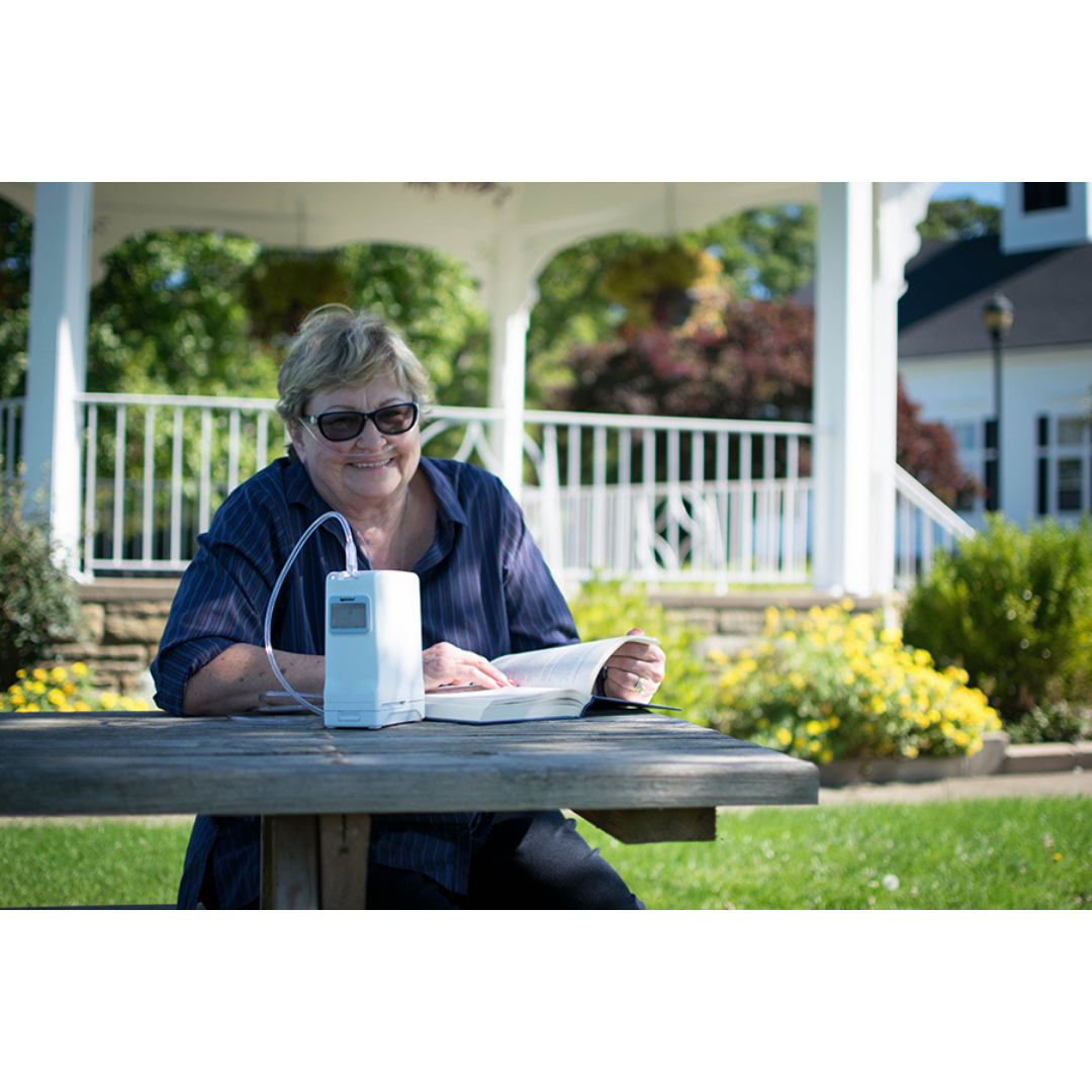An older woman wearing sunglasses sits at a picnic table outdoors, smiling and reading a book beside her OxyGo FIT Compact Portable Oxygen Concentrator. A white gazebo and garden are visible in the background.