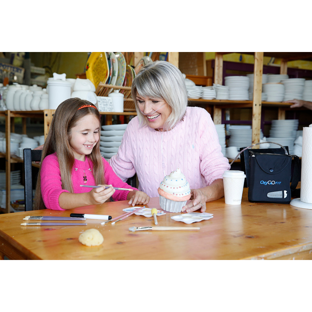 An older woman using an OxyGo FIT Compact Portable Oxygen Concentrator and a young girl smile while decorating a ceramic cupcake at a table with paintbrushes and art supplies in a pottery studio, with shelves of white ceramics in the background.