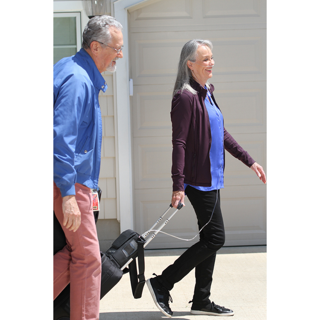An older woman, casually dressed, smiles while walking outside with a black rolling suitcase and her OxyGo NEXT Ultralite Portable Oxygen Concentrator, beside an older man, as they pass a beige garage door on a sunny day.