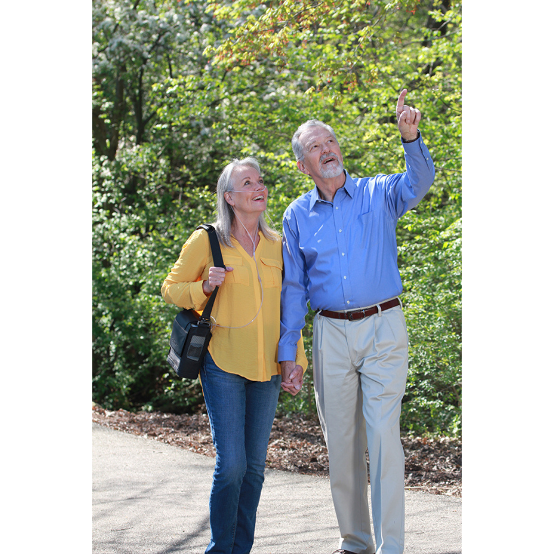 An older couple strolls hand in hand on a sunny day, smiling and relaxed amid green trees, enjoying the freedom provided by their OxyGo NEXT Ulralite Portable Oxygen Concentrator.