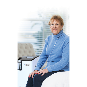 An older woman in a blue sweater sits on a bed, smiling, using a nasal oxygen tube connected to an OxyGo OxyHome 5L Stationary Concentrator beside her in a bright room with large windows and blinds.