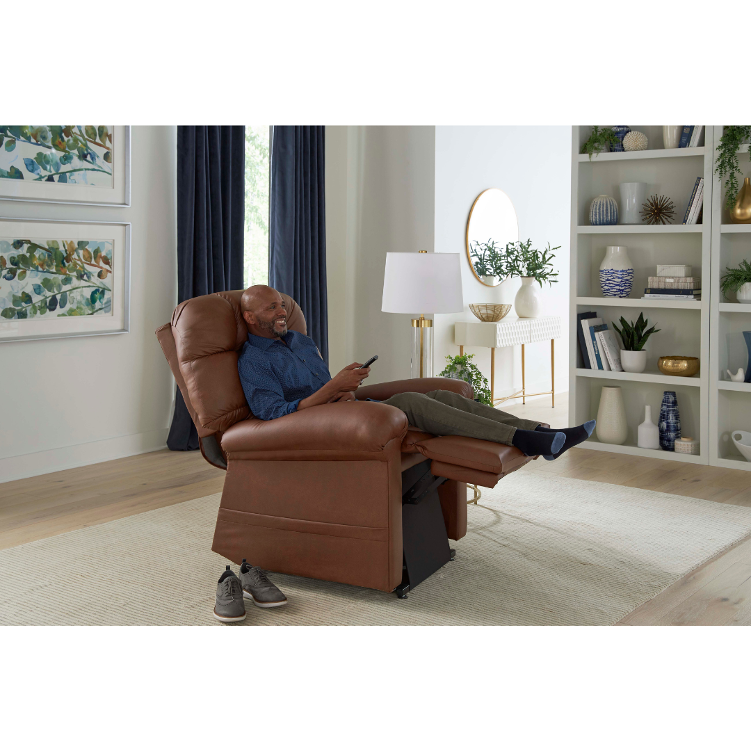 A man relaxes with his feet up and a remote in hand, smiling in a bright living room, in the Golden Tech PR515 Cloud Recliner With Lift Assist, ZG+ & Twilight. Shoes are on the floor beside the chair, surrounded by a bookshelf and lamp.
