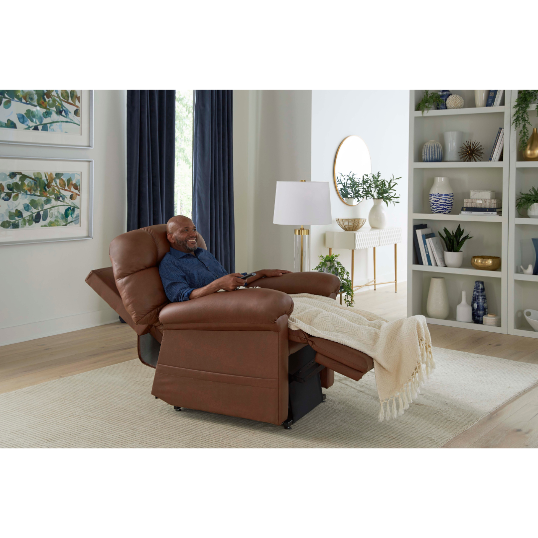 A man relaxes in the Golden Tech PR515 Cloud Recliner With Lift Assist, ZG+ & Twilight, his legs elevated under a light blanket in a bright, modern living room adorned with plants, artwork, and shelves.