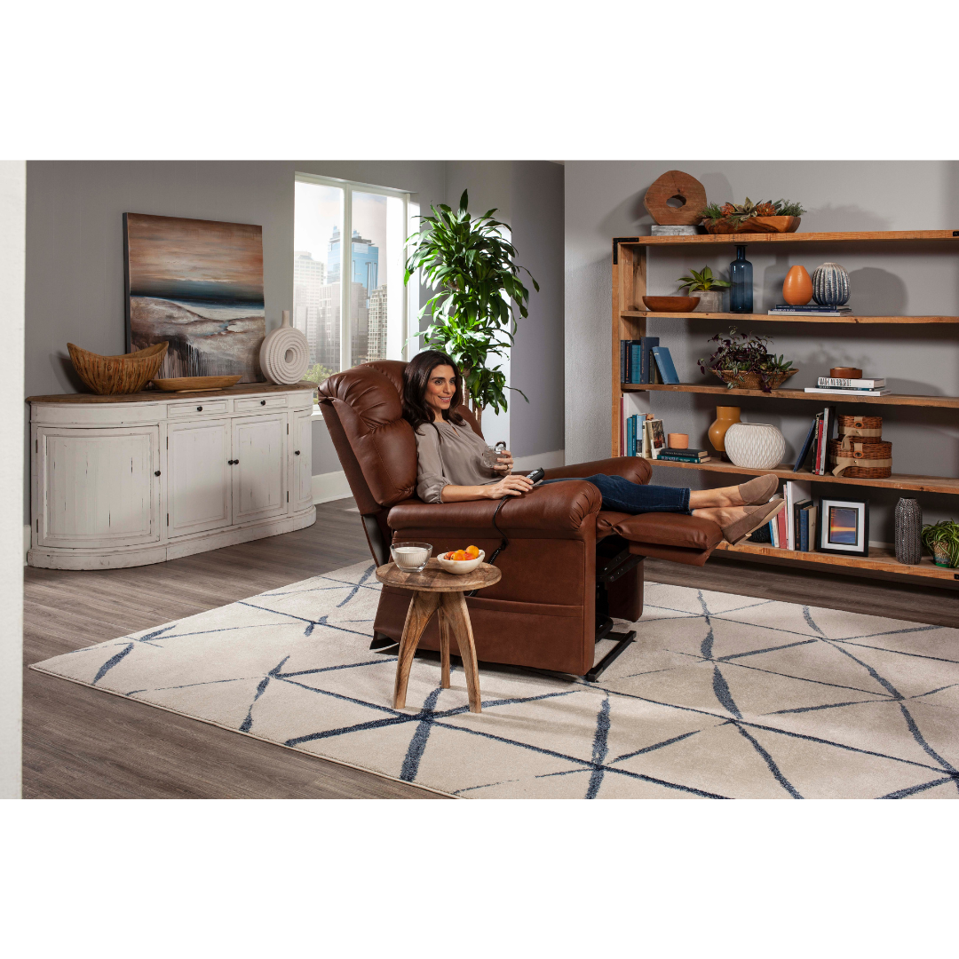 A person relaxes on a Golden Tech PR515 Cloud Recliner with Lift Assist, feet up, enjoying ergonomic support in a cozy living room. They hold a remote as the room features a large bookshelf with decor, white sideboard, potted plant, and modern wavy-lined rug on wooden flooring.