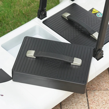 A close-up of a black rectangular foot pedal with a handle, attached to the Paiseec Mobility Scooter D3 Battery on a white outdoor surface. Grass and paving stones can be seen in the background.