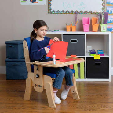 A young girl sits in a Circle Specialty Pango Pediatric Activity Chair at a desk, cutting red paper with scissors. A bottle of glue is nearby, with colorful storage bins and classroom decorations in the background.