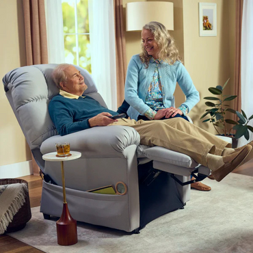 An elderly man relaxes in his Journey Perfect Sleep Reclining Lift Chair with Heat, remote in hand. A smiling gray-haired woman stands beside him. The chair provides therapeutic heat and massage for top-notch comfort. A small table holds a drink as soft natural light brightens the room.