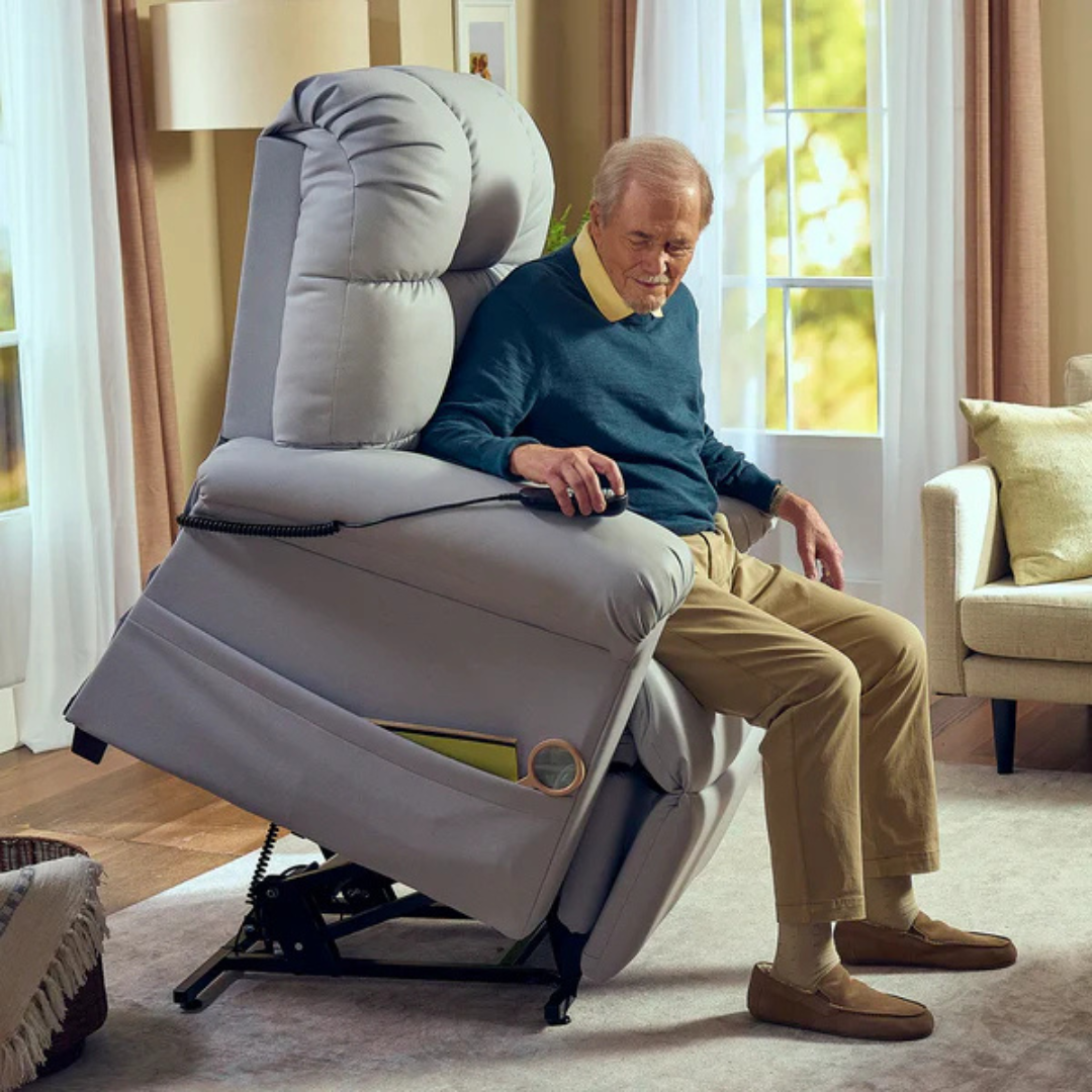An elderly man sits on a Journey Perfect Sleep Reclining Lift Chair with Heat, tilted forward to help him stand. Dressed in a blue sweater and beige pants, he relishes the comfort of the chair in a cozy room with curtained windows and a beige sofa.