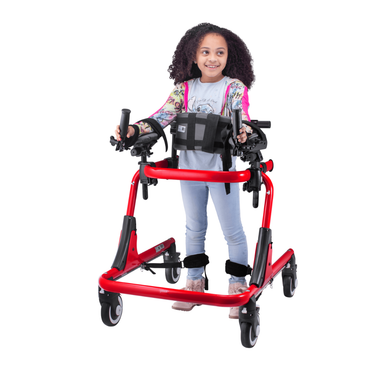 A young girl with curly hair smiles and looks to the side while using the Circle Specialty Pediatric Pivot Gait Trainer, a bright red adjustable walker with arm supports and wheels. She wears light blue pants and a colorful long-sleeve shirt.