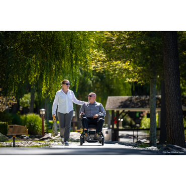 A man in a Pride Mobility Jazzy® Ultra Light Portable Electric Wheelchair, featuring a sleek carbon fiber frame, navigates a serene park path. A woman, his travel companion with a shopping bag, walks beside him as they converse amidst lush greenery and a small pavilion.