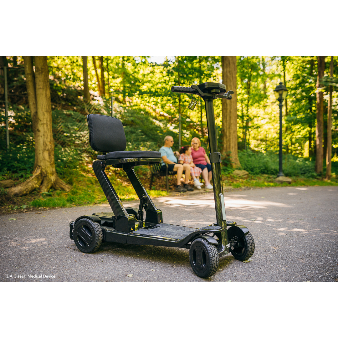 A sleek Pride Mobility GoGo Carbon Ultralite Scooter in black, weighing only 35 lbs, is parked on a paved path in a lush park. In the background, three people slightly out of focus sit on a bench enjoying the natural surroundings on a sunny day.