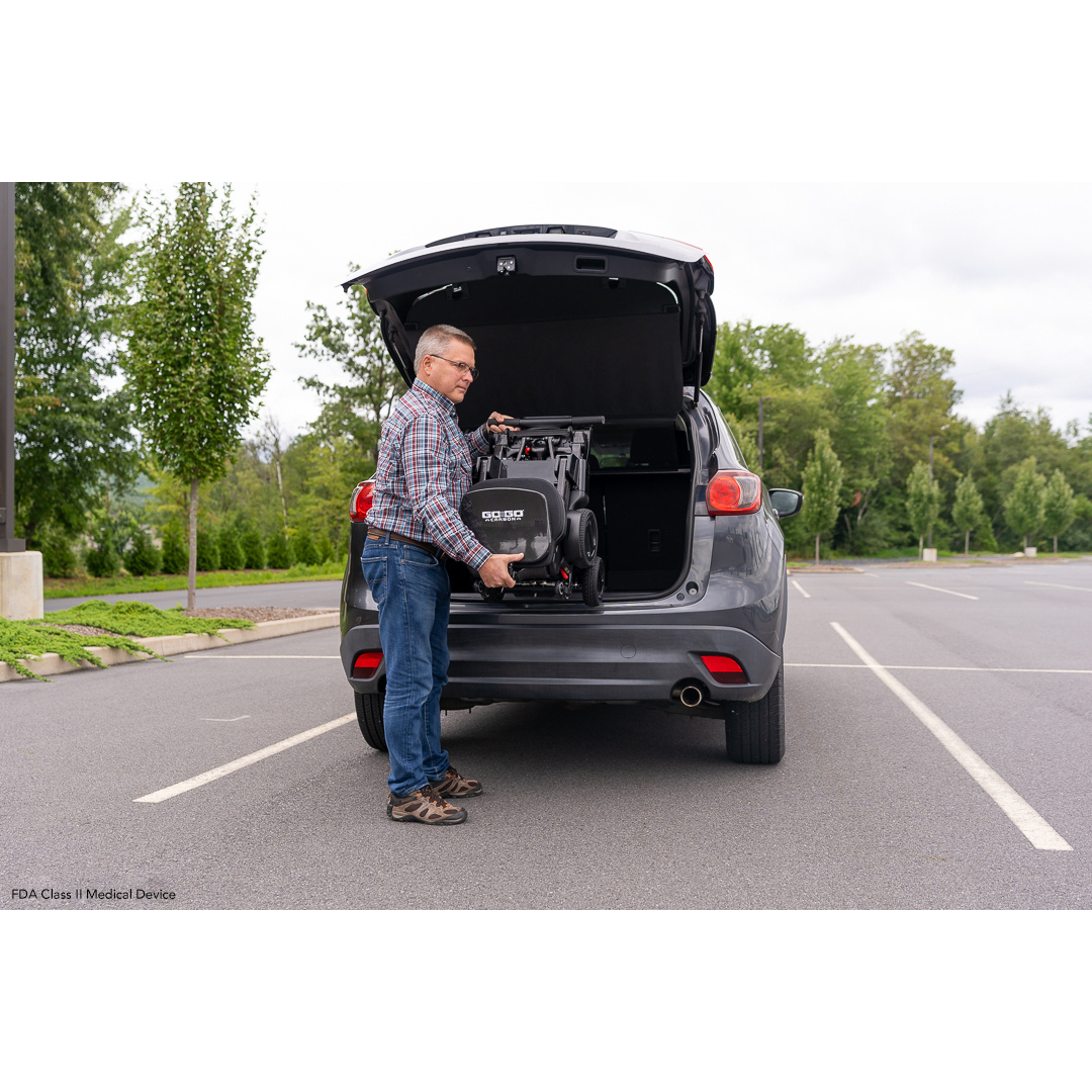 In a parking lot, a man is loading a portable lift device and the Pride Mobility GoGo Carbon Ultralite Mobility Scooter, weighing just 35 lbs, into the trunk of an SUV parked on an empty road. The vehicle is surrounded by swaying grass and towering trees beneath an overcast sky.