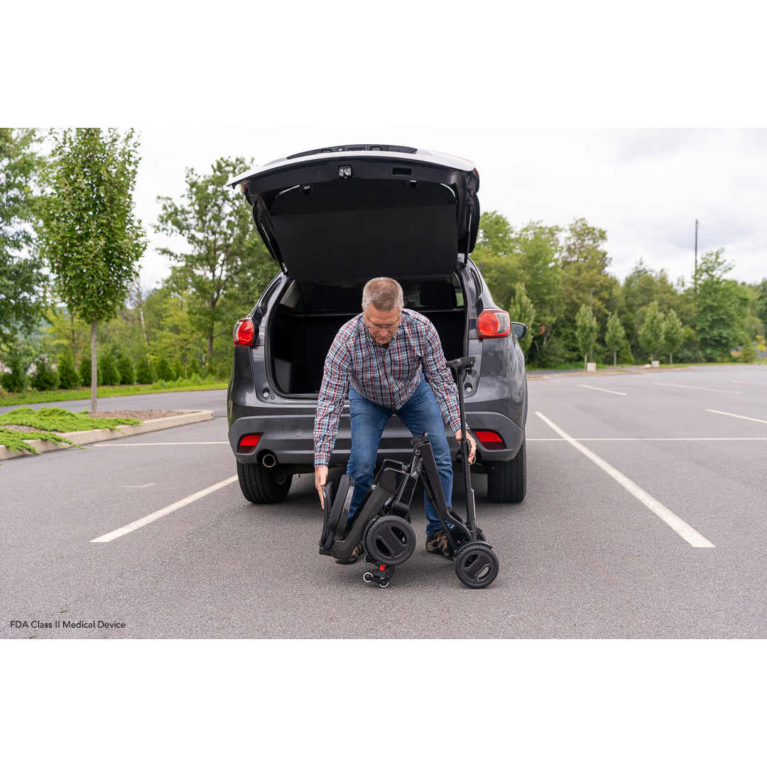 A man in a plaid shirt unloads his Pride Mobility GoGo Carbon Ultralite Scooter, weighing only 35 lbs, from the trunk of a gray SUV in an empty parking lot. With trees and clouds framing the scene, he appreciates its lightweight design for easy transport.