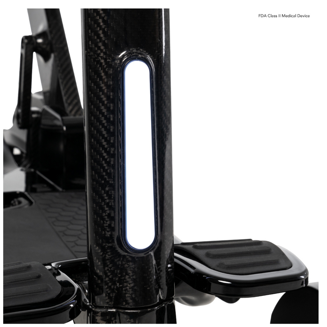 Close-up of the black carbon fiber seat post with step supports from the Pride Mobility GoGo Carbon Ultralite Mobility Scooter (35 lbs). A white rectangular light panel is embedded, and FDA Class II Medical Device text emphasizes its portable design.