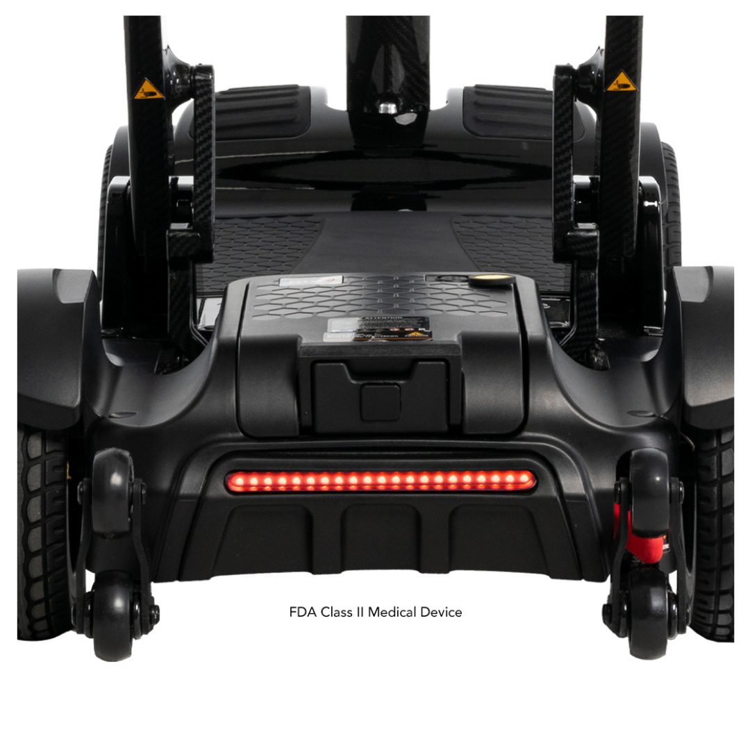 Close-up of the sleek black base of the Pride Mobility GoGo Carbon Ultralite Scooter, featuring a textured footrest, durable wheels, and a glowing red rear light on its carbon fiber frame. Text reads: FDA Class II Medical Device.
