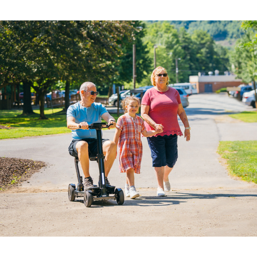 A man on a Pride Mobility GoGo Carbon Ultralite Scooter (only 35 lbs), a young girl in a plaid dress, and a woman in a pink shirt enjoy walking together on a sunny path, with trees and buildings providing the perfect backdrop for their leisurely outing.