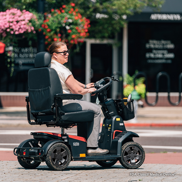 A woman in sunglasses rides a Pride Mobility PX4 HD Bariatric Scooter on a city sidewalk, with flowers and buildings in the background.