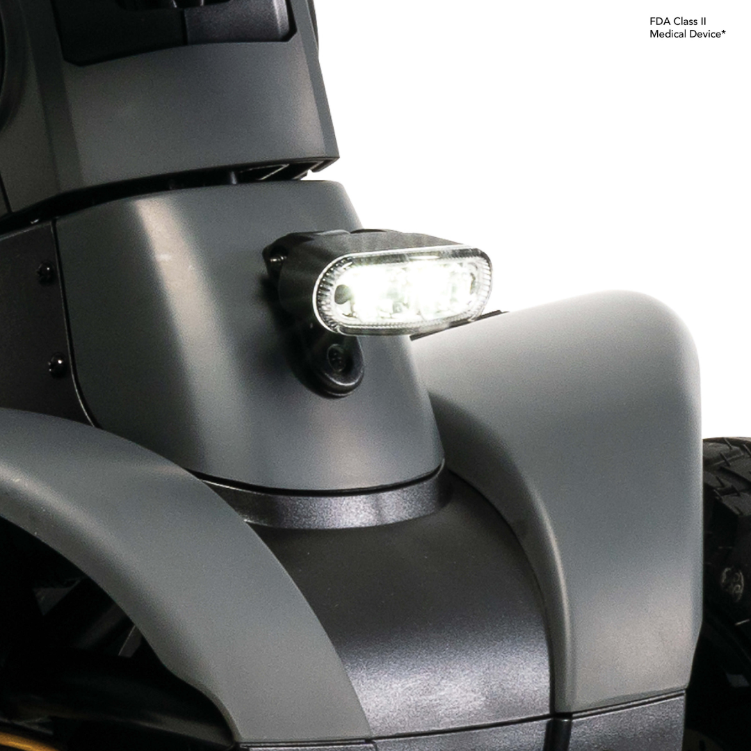 Close-up of the Pride Mobility Pursuit® 2 Offroad HD Scooter’s front, featuring a durable matte gray body, bright LED headlight above the wheel, and part of a black tire.