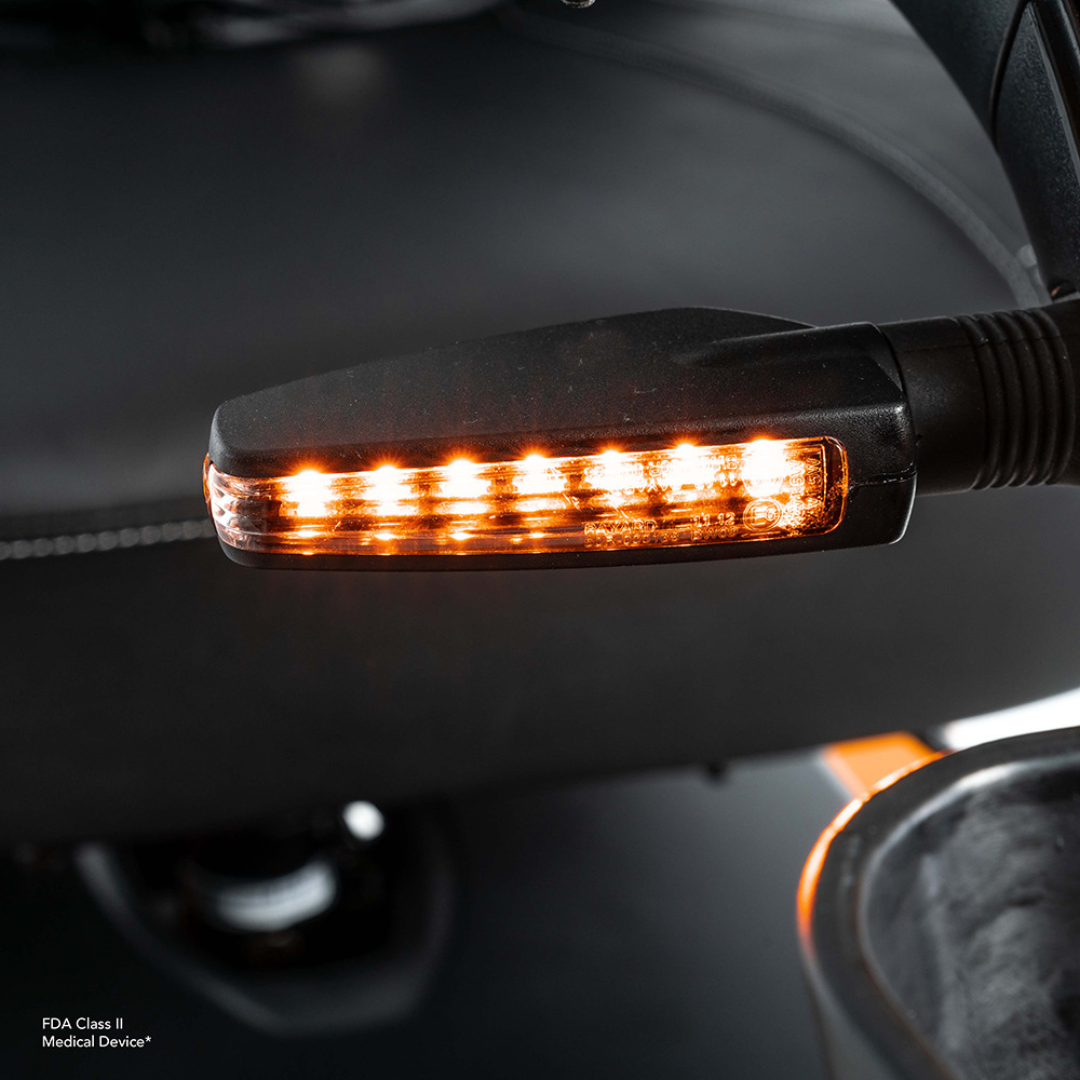 Close-up of the illuminated amber turn signal on the side mirror of the Pride Mobility Pursuit® 2 Offroad HD Scooter, featuring multiple small LED bulbs in a row, set against a blurred dark background.