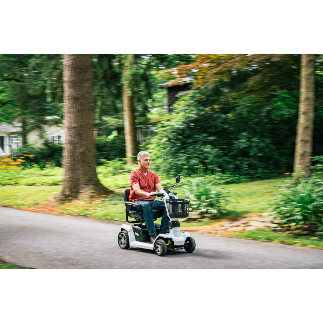 An older man in a red shirt rides a Pride Mobility Zero Turn 10 HD Scooter along a paved path in a green, tree-filled park, the blurred background emphasizing motion.