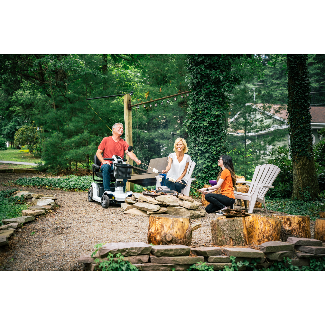 Three adults chat by a stone fire pit in a wooded backyard. One man relaxes on his Pride Mobility Zero Turn 10 HD Scooter featuring EZ Turn Technology, while two women sit nearby, surrounded by lush greenery.