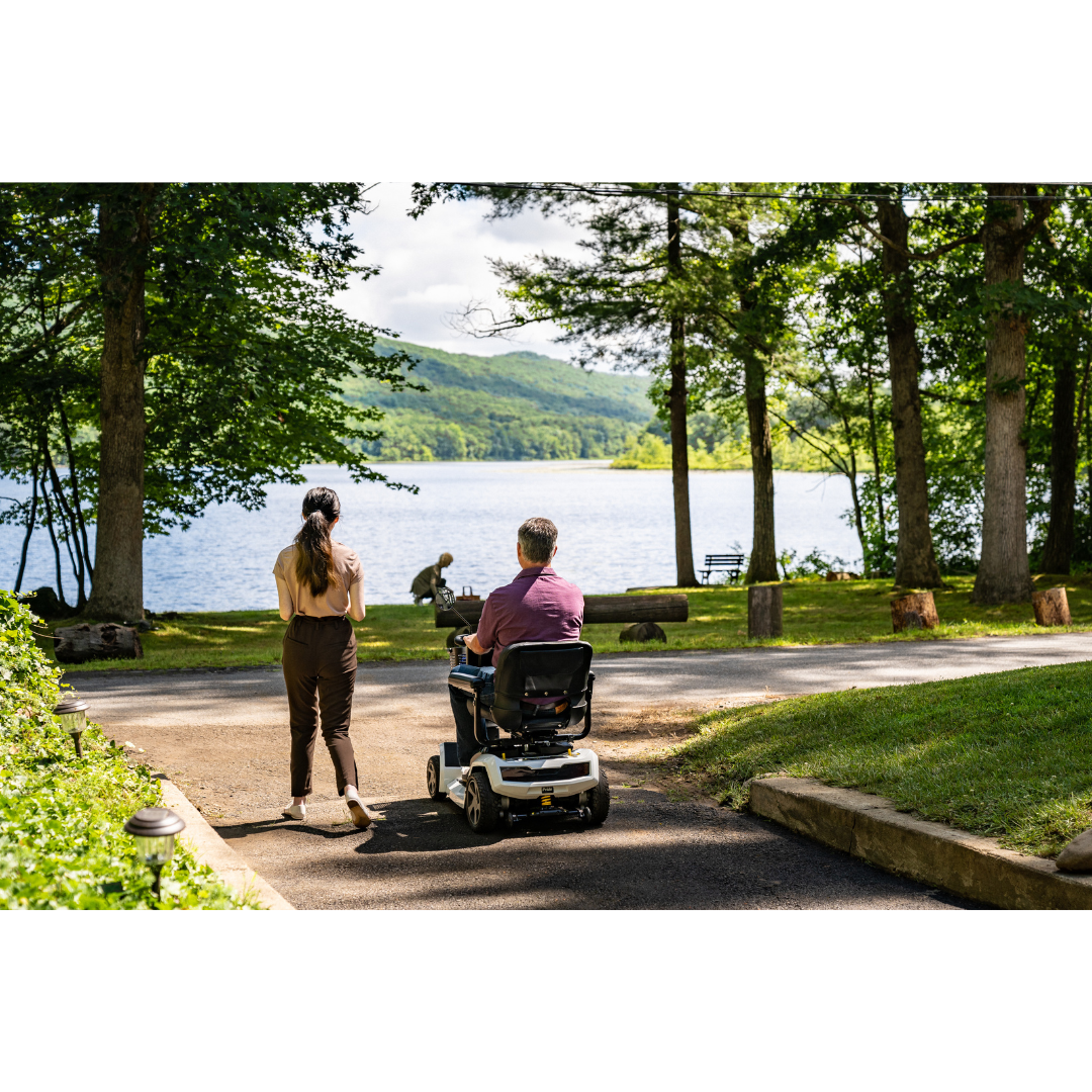 A woman walks beside a man riding a Pride Mobility Zero Turn 10 HD Scooter with EZ Turn Technology along a lakeside path, surrounded by trees, greenery, and mountains in the distance.