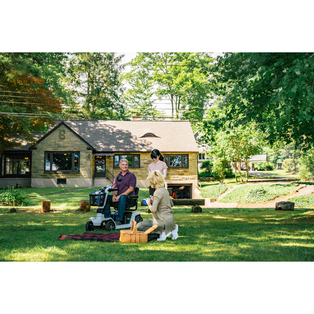 Three people have a picnic on a grassy lawn by a house; a man relaxes in his Pride Mobility Zero Turn 10 HD Scooter while two women kneel on a blanket with a picnic basket under the shade of trees.