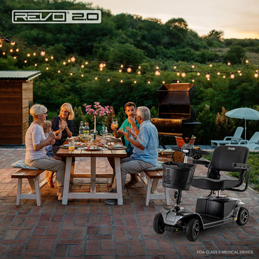 Four older adults laugh and share a meal at a picnic table outdoors at sunset. String lights glow above, while a Pride Mobility Revo® 2.0 4-Wheel Scooter rests nearby amid lush greenery, creating a warm and inviting patio setting.
