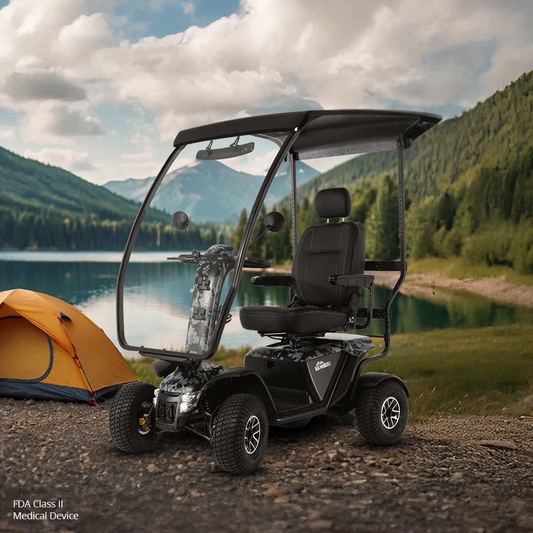 A Pride Baja® Wrangler® 2 Heavy Duty Outdoor Scooter with Canopy, black with four wheels, is parked on dirt by a lake near a yellow tent. Forested hills and a cloudy sky highlight its all-terrain outdoor performance.