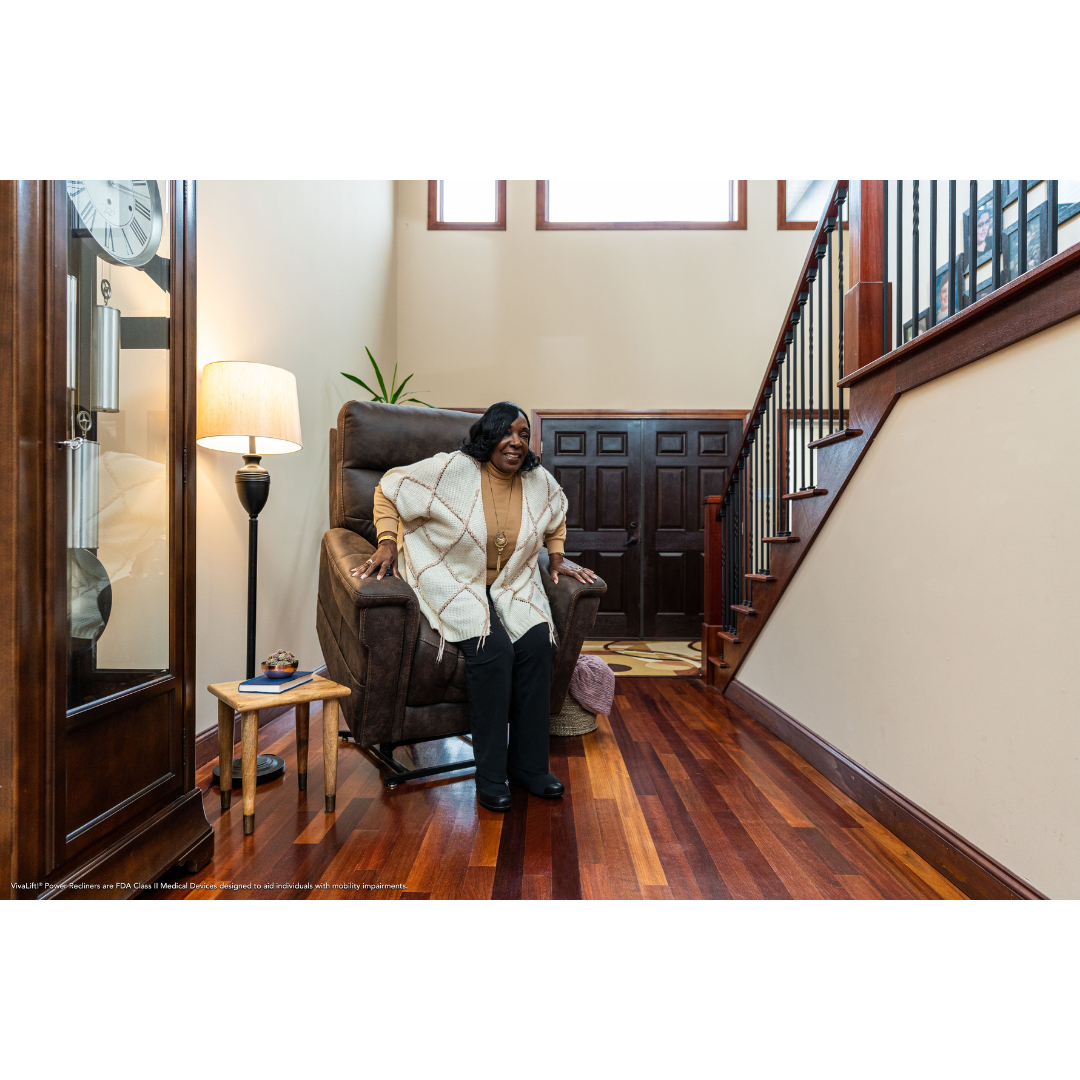 An older woman relaxes on a Pride Mobility VivaLift! Radiance Recliner with Lift Assist & Heat in a bright living room with wooden floors, staircase, lamp, side table, and large windows letting in natural light above.