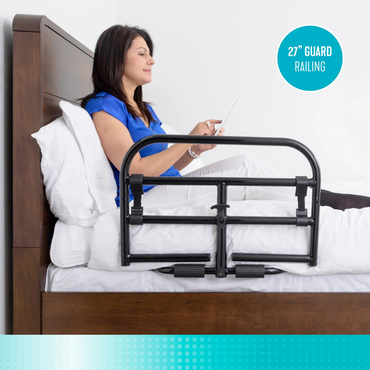 A woman sits upright in bed using a tablet, with a black Stander Prime Bariatric Safety Bed Rail attached to the side of the bed for added bedside safety.